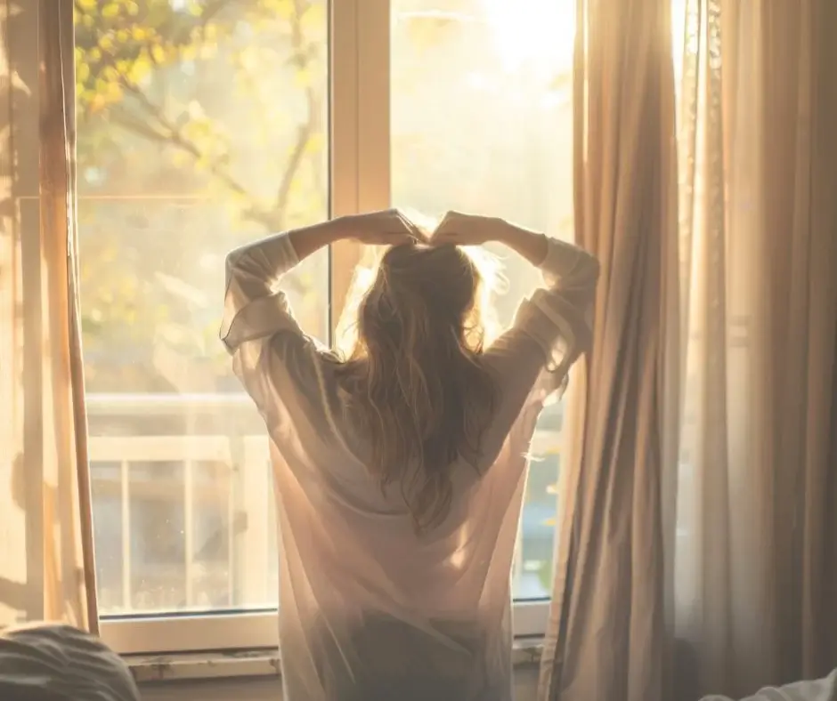 Woman standing by a window in morning light, stretching and greeting the new day with calm.