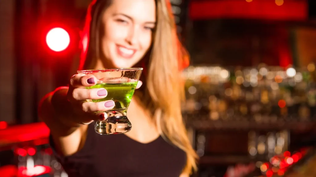 Kobieta w barze trzymająca kieliszek z zielonym napojem w wyciągniętej dłoni – moment oczekiwania i pierwszego wrażenia smaku.