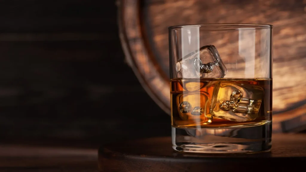 A glass of amber-colored whisky-like drink with ice cubes on a wooden surface, with a barrel blurred in the background.