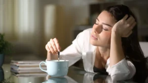 Tired woman sitting at a table, stirring a drink in a mug and looking low on energy.