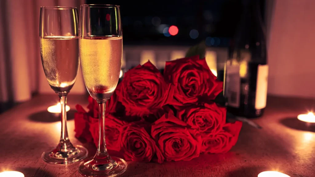 Two glasses with a light non-alcoholic drink on a table, next to red roses and candles in a romantic Valentine’s Day evening setting.