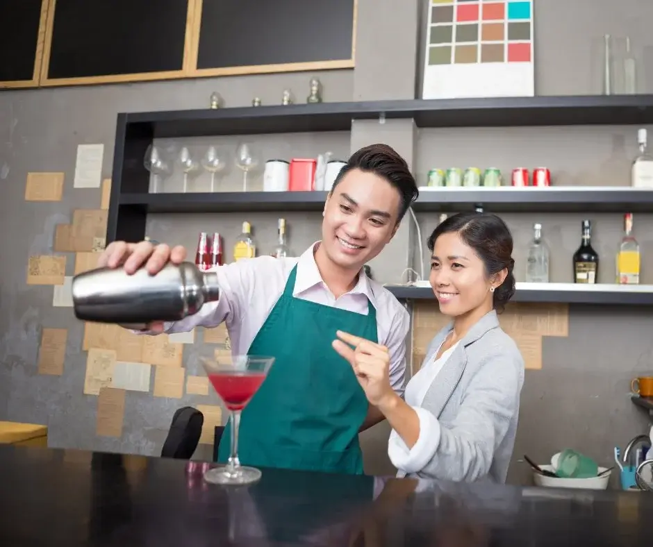 Two people preparing a non-alcoholic drink at a bar, one pouring the drink into a glass while the other watches and smiles.