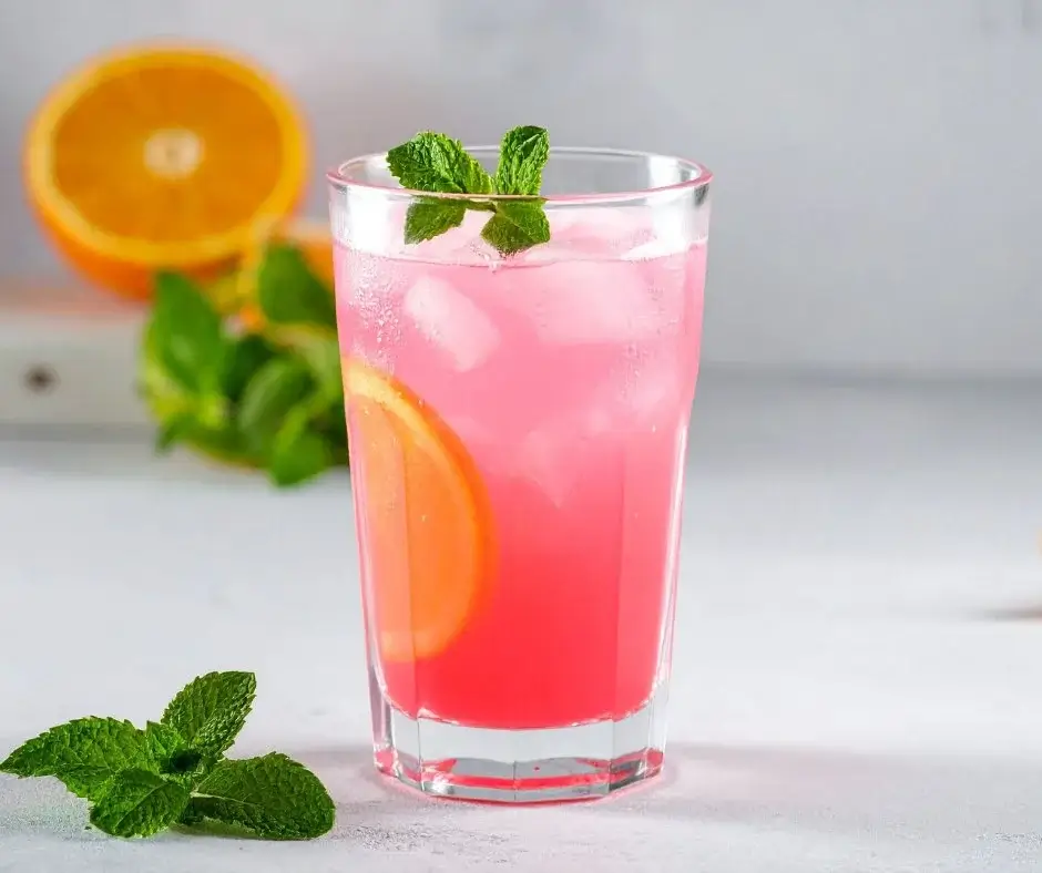 Różowy bezalkoholowy drink z lodem, plasterkiem pomarańczy i listkami mięty w szklance na jasnym tle.